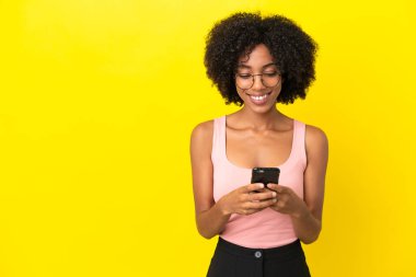 Sarı arka planda izole edilmiş genç Afrikalı Amerikalı kadın cep telefonuyla mesaj gönderiyor.