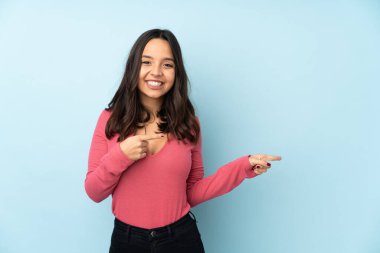 Mavi arka planda izole edilmiş genç melez kadın şaşırmış ve işaret ediyor.