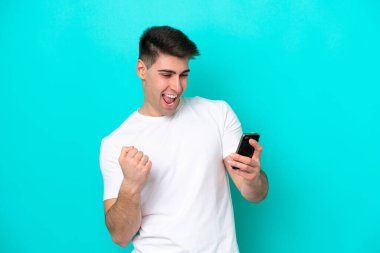 Genç beyaz adam mavi arka planda izole edilmiş cep telefonu kullanıyor ve zafer jesti yapıyor.