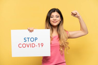 Sarı arka planda izole edilmiş genç bir kız elinde yazılı bir pankartla Covid 19 'u durdur ve güçlü bir hareket yap.