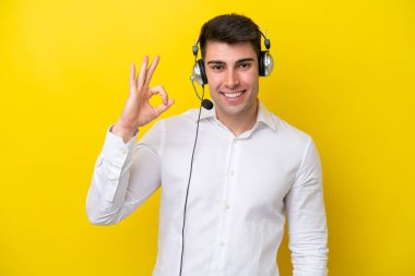 Tele pazarlamacı beyaz adam sarı arka planda izole edilmiş bir kulaklıkla çalışıyor. Parmaklarıyla tamam işareti gösteriyor.