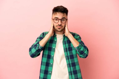 Pembe arka planda izole edilmiş genç beyaz adam hayal kırıklığına uğramış ve kulaklarını kapatmış.