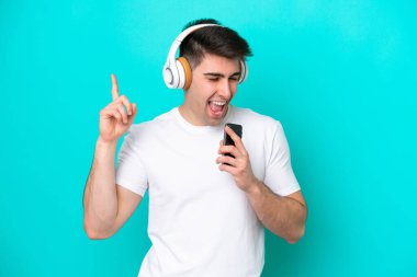Kafkasyalı genç adam mavi arka planda izole edilmiş. Cep telefonuyla müzik dinliyor ve şarkı söylüyor.