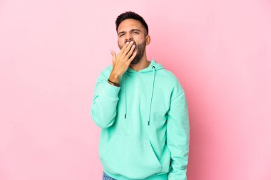 Pembe arka planda izole edilmiş genç beyaz adam esniyor ve ağzını eliyle açık tutuyor.