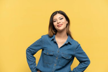 Sarı arka planda izole edilmiş genç bir kız kolları kalçasında ve gülümsüyor.