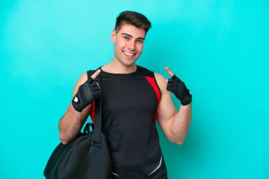 Mavi arka planda izole edilmiş spor çantalı beyaz bir genç baş parmak hareketi yapıyor.