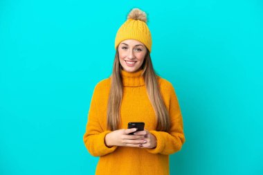 Kışlık ceket giyen genç beyaz kadın mavi arka planda izole edilmiş cep telefonuyla bir mesaj gönderiyor.