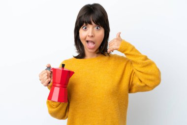 Beyaz arka planda kahve demliği tutan melez bir kadın telefon jesti yapıyor. Bana geri dönüş işareti ver.