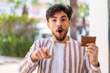Açık havada cüzdan tutan yakışıklı bir Arap şaşırmış ve ön tarafı işaret ediyor.