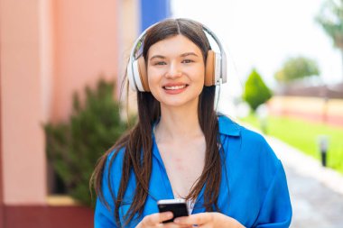 Genç ve güzel bir kadın açık havada müzik dinliyor cep telefonu ve ön tarafa bakıyor.