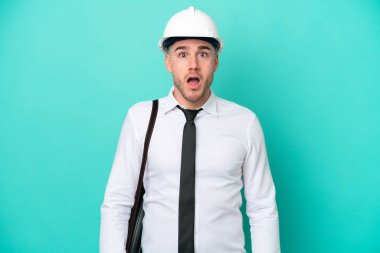 Miğferli ve elinde mavi arka planda izole edilmiş planları olan genç bir beyaz adam yukarı bakıyor ve şaşırmış bir ifadeyle