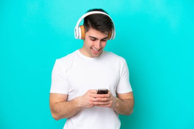 Kafkasyalı genç adam mavi arka planda izole edilmiş müzik dinliyor ve cep telefonu arıyor.