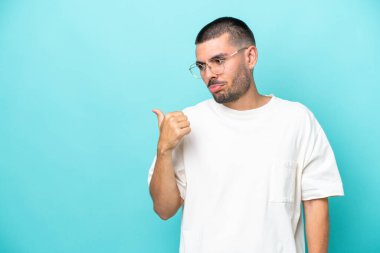 Young caucasian man isolated on blue background unhappy and pointing to the side