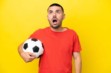Futbol oynayan genç beyaz adam sarı arka planda izole edilmiş yukarı bakıyor ve şaşırmış bir ifadeyle