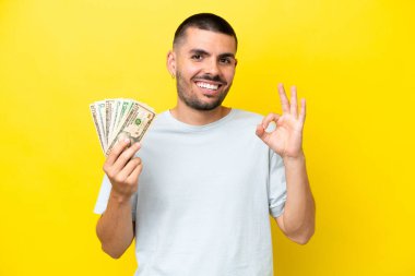 Kafkasyalı genç adam çok para alıyor. Sarı arka planda izole edilmiş. Parmaklarıyla işaret veriyor.
