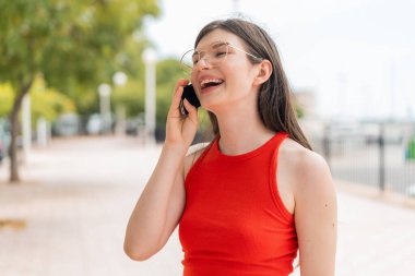 Dışarıda gözlüklü genç bir kadın cep telefonuyla sohbet ediyor.