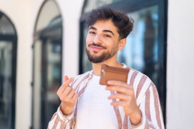Dışarıdaki yakışıklı bir Arap, elinde para dolu bir cüzdan tutuyor.