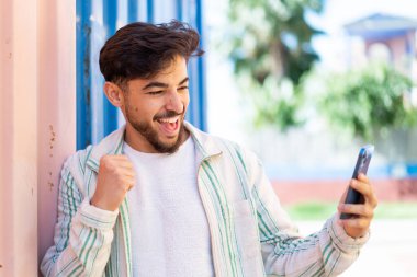 Yakışıklı bir Arap açık havada cep telefonu kullanıyor ve zaferi kutluyor.