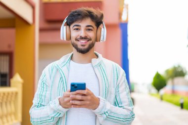 Yakışıklı bir Arap açık havada cep telefonuyla müzik dinliyor ve önüne bakıyor.