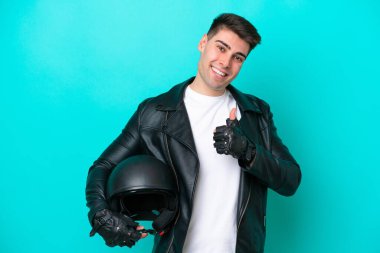 Motosiklet kasklı genç beyaz adam mavi arka planda izole edilmiş bir şekilde baş parmak işareti yapıyor.