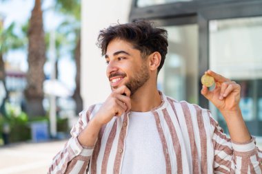 Yakışıklı bir Arap 'ın elinde Bitcoin tutarken bir fikir üretip yan gözle bakması.