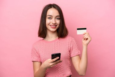 Pembe arka planda izole edilmiş genç Ukraynalı bir kadın kredi kartıyla cep telefonuyla alışveriş yapıyor.