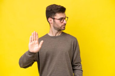 Sarı arka planda izole edilmiş genç beyaz adam dur hareketi yapıyor ve hayal kırıklığına uğruyor.