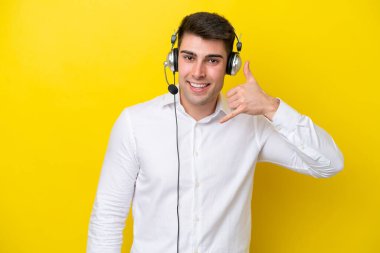 Tele pazarlamacı beyaz adam sarı arka planda izole edilmiş bir kulaklıkla çalışıyor ve telefon jesti yapıyor. Bana geri dönüş işareti ver.