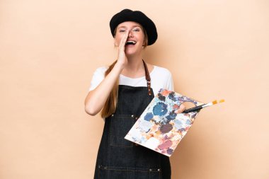 Beyaz tenli genç bir kadın bej arka planda izole edilmiş bir palet tutuyor ve ağzı açık bağırıyor.