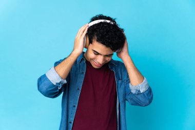 Genç Venezuelalı adam mavi arka planda izole edilmiş müzik dinliyor.