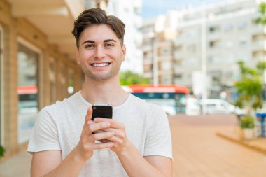 Genç yakışıklı adam kameraya bakıyor ve cep telefonunu kullanırken gülümsüyor.