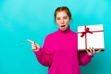 Kafkasyalı genç bir kadın mavi arka planda izole edilmiş bir hediye tutuyor. Şaşırmış ve işaret ediyor.