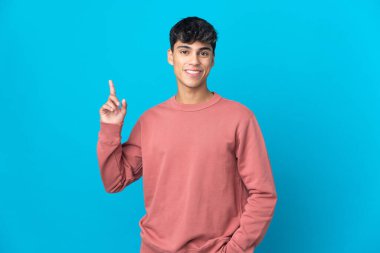İzole edilmiş mavi arka planı olan genç adam en iyinin işareti olarak parmağını kaldırıyor ve gösteriyor.