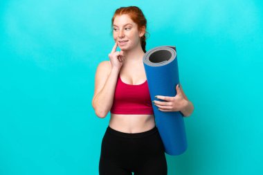 Genç spor kırmızısı kadın yoga derslerine giderken elinde mavi arka planda bir minder tutarken yukarı bakarken bir fikir düşünüyor.