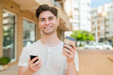 Genç yakışıklı adam cep telefonu kullanıyor ve elinde mutlu bir ifadeyle kahve tutuyor.