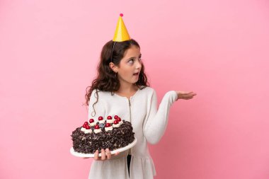Küçük beyaz kız elinde doğum günü pastası tutuyordu. Pembe arka plandaydı ve yüzünde sürpriz bir ifade vardı.