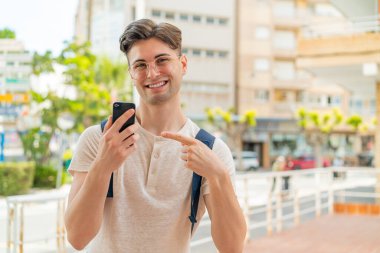 Genç ve yakışıklı bir adam açık havada cep telefonu kullanıyor ve onu işaret ediyor.