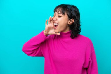 Arjantinli genç kadın mavi arka planda izole edilmiş ve ağzı sonuna kadar açık bağırıyor.