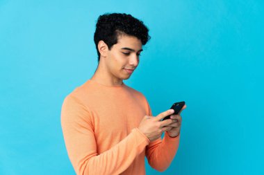 Venezuelalı adam mavi arkaplanda izole edilmiş cep telefonuyla bir mesaj ya da e-posta gönderiyor