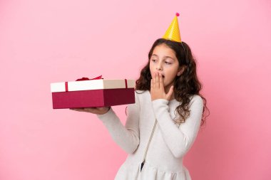 Küçük beyaz kız pembe arka planda saklanmış bir hediye tutuyor. Şaşırmış ve şok olmuş bir yüz ifadesi var.