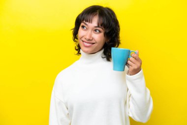 Arjantinli genç bir kadın elinde kahve fincanı tutarken sarı arka planda izole edilmiş gülümserken yukarı bakıyor.