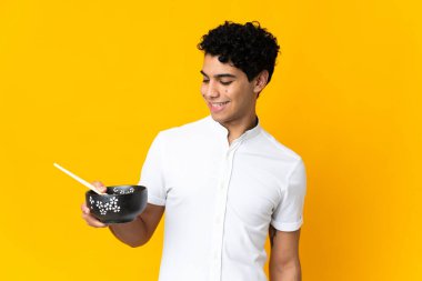 Genç Venezuelalı adam, elinde çubuklarla elinde bir kase şehriye tutarken mutlu bir ifadeyle sarı arka planda izole edilmiş.