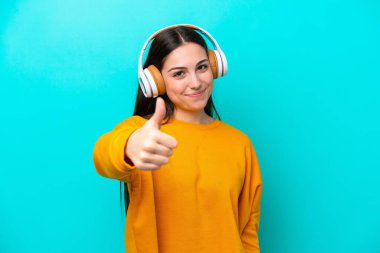 Mavi arka planda izole edilmiş genç bir kız müzik dinliyor ve başparmağını kaldırıyor.