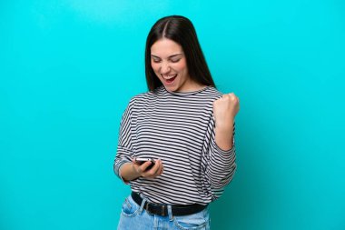 Genç beyaz kadın mavi arka planda izole edilmiş ve telefon zafer pozisyonunda.
