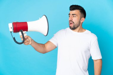 Caucasian man over isolated blue background holding a megaphone with stressed expression