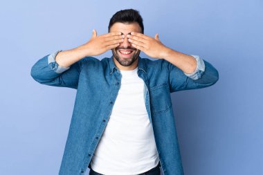 İzole edilmiş mavi arka plan üzerinde beyaz bir adam. Gözleri elleriyle kaplıyor ve gülümsüyor.