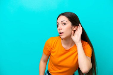 Mavi arka planda izole edilmiş genç beyaz kadın elini kulağına koyarak bir şeyler dinliyor.