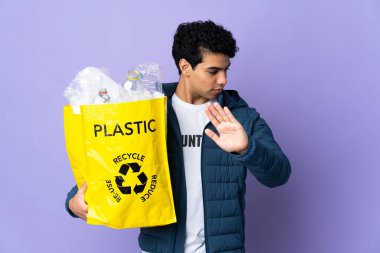 Genç Venezuelalı bir adam elinde bir çanta dolusu plastik şişeyle durup jest yapıyor ve hayal kırıklığına uğruyor.