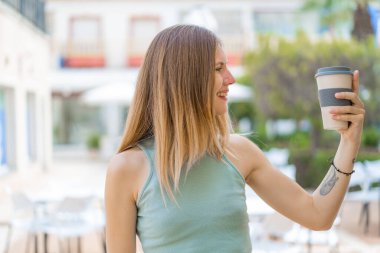 Genç sarışın kadın açık havada kahveyi elinde tutuyor, mutlu bir ifadeyle
