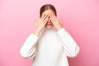 Pembe arka planda izole edilmiş genç beyaz kadın gözlerini elleriyle kapatıyor.